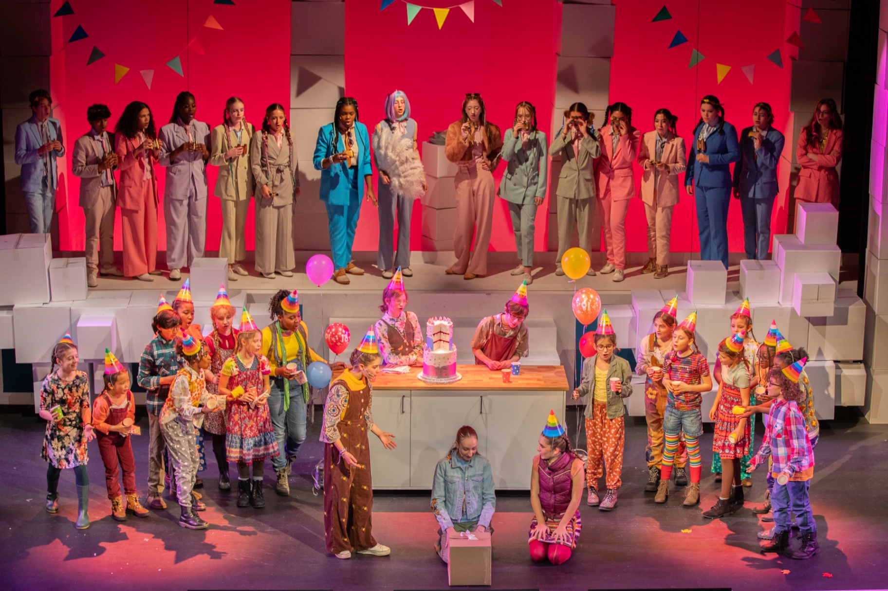 The full company of Pandora's Box on stage against a bold pink backdrop, the mortals gathered around Pandora as she opens her gift.