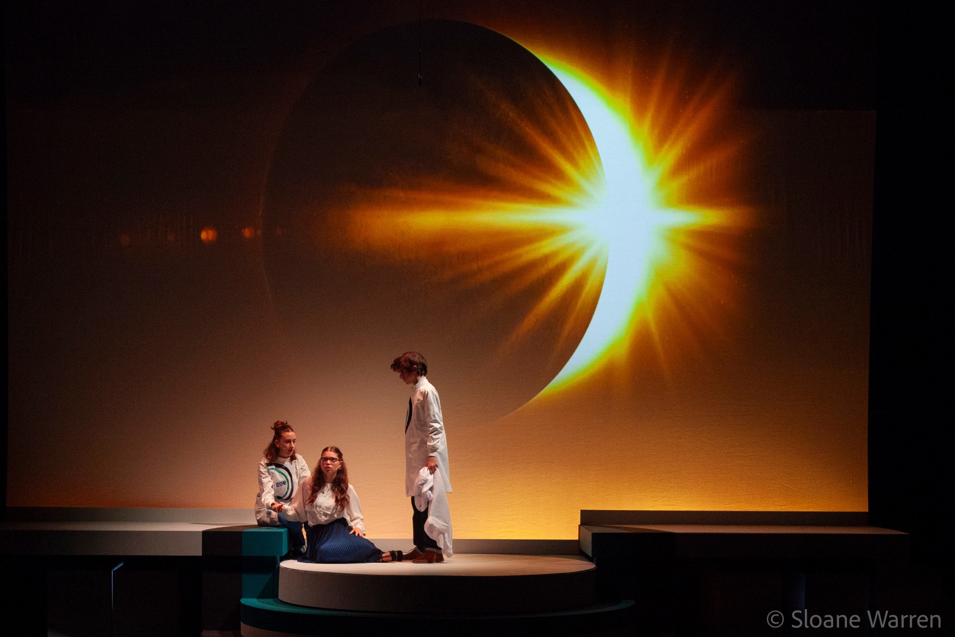 Three young scientists in white lab coats stand before a vast projected solar eclipse in The Quest.